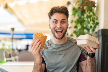 Young handsome man taking fried chips and takeaway box food with surprised expression