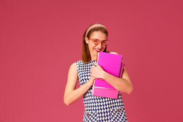 Beautiful, smiling young woman in pink sunglasses standing with notebooks against pink studio background. Freelancer. Concept of youth, human emotions, education, lifestyle, fashion