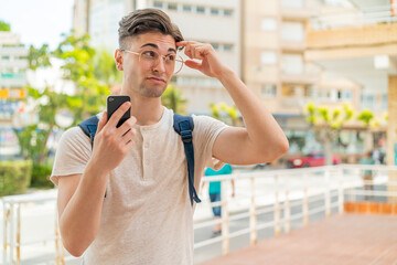Young handsome man using mobile phone at outdoors having doubts and with confuse face expression