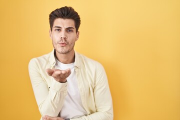 Young hispanic man standing over yellow background looking at the camera blowing a kiss with hand on air being lovely and sexy. love expression.