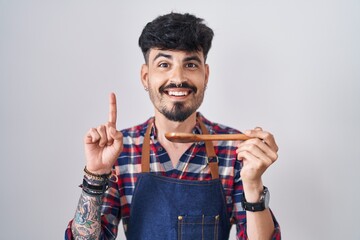 Young hispanic man with beard wearing apron tasting food holding wooden spoon surprised with an idea or question pointing finger with happy face, number one