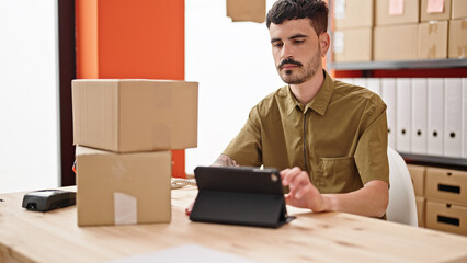 Young hispanic man ecommerce business worker holding packages using touchpad at office