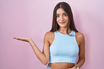 Young brunette woman standing over pink background smiling cheerful presenting and pointing with palm of hand looking at the camera.