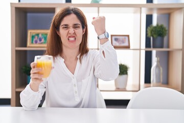 Brunette woman drinking glass of orange juice angry and mad raising fist frustrated and furious while shouting with anger. rage and aggressive concept.