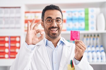 Hispanic man with beard working at pharmacy drugstore holding condom doing ok sign with fingers, smiling friendly gesturing excellent symbol