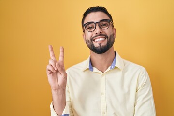 Hispanic young man wearing business clothes and glasses smiling with happy face winking at the camera doing victory sign with fingers. number two. - obrazy, fototapety, plakaty