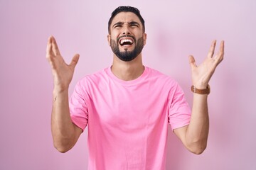 Hispanic young man standing over pink background crazy and mad shouting and yelling with aggressive expression and arms raised. frustration concept. - obrazy, fototapety, plakaty