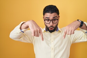 Hispanic young man wearing business clothes and glasses pointing down with fingers showing advertisement, surprised face and open mouth