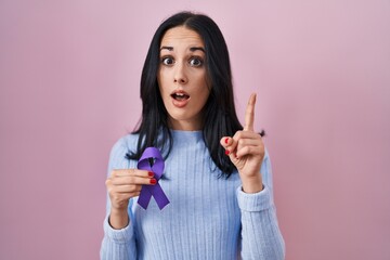 Hispanic woman holding purple ribbon awareness surprised with an idea or question pointing finger...