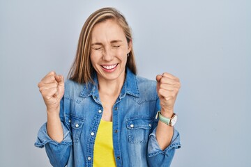 Young blonde woman standing over blue background excited for success with arms raised and eyes...