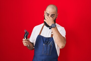 Young hispanic man with beard and tattoos wearing barber apron holding razor smelling something...