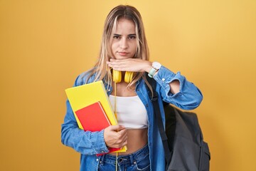 Young blonde woman wearing student backpack and holding books cutting throat with hand as knife,...