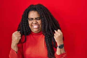 Plus size hispanic woman standing over red background very happy and excited doing winner gesture with arms raised, smiling and screaming for success. celebration concept.