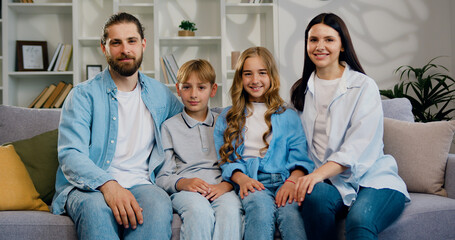 Family portrait of father, mother and two lovely children smiling at camera and hugging in modern living room at home. Good looking caucasian woman and man witch son and daughter sitting on the sofa