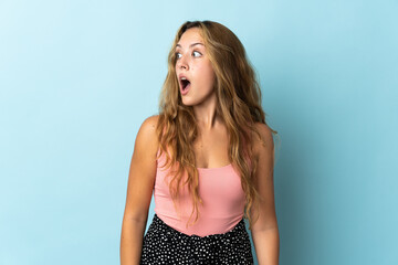 Young blonde woman isolated on blue background doing surprise gesture while looking to the side