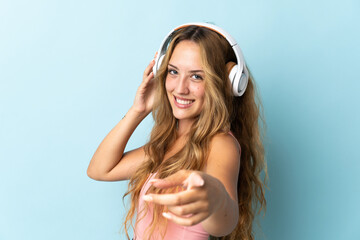 Young blonde woman isolated on blue background listening music and pointing to the front - obrazy, fototapety, plakaty