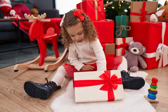 Adorable blonde girl unpacking gift sitting by christmas tree at home
