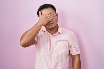 Chinese young man standing over pink background covering eyes with hand, looking serious and sad....