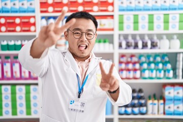 Chinese young man working at pharmacy drugstore smiling with tongue out showing fingers of both...