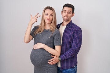 Young couple expecting a baby standing over white background shooting and killing oneself pointing...