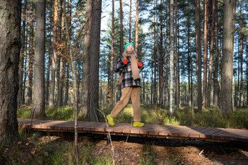 Woman middle aged tourist walking resting on eco trail in Scandinavian pine forest along wooden path. Relaxed female enjoying pastime in unity with nature. Travel tourism wanderlust vacation concept.