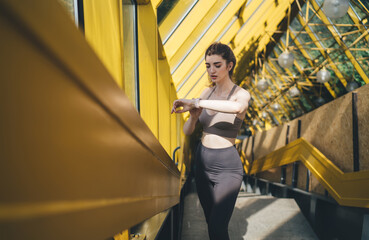 Focused young Caucasian woman in a taupe sports top and black leggings adjusting her fitness watch...