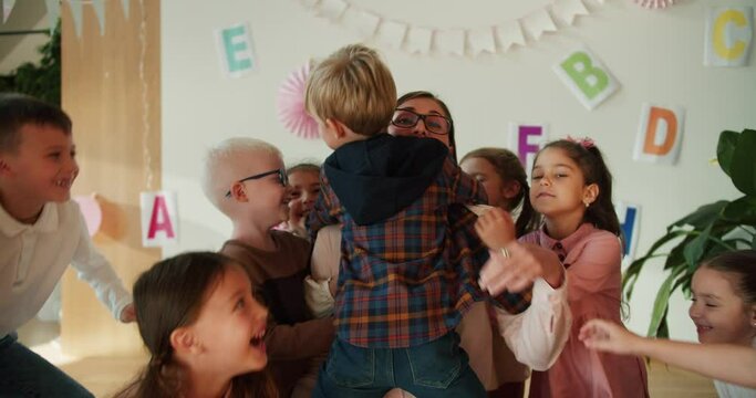 a group of children hug their teacher, a blonde woman with a bob hairstyle in glasses in a white shirt at their first lesson in a club for preparing children for school classes. The children love