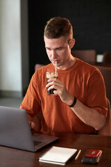 Positive young man enjoying cold soft drink when reviewing programming code on laptop