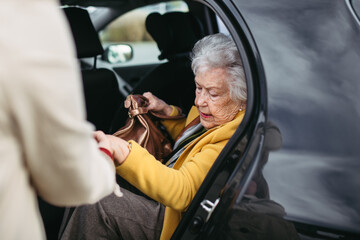 Senior lady getting out of the car, caregiver helping her, holding her hands. Elderly woman has...