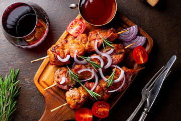 Pork kebab with barbecue sauce and tomatoes and a glass of red wine on a dark background, top view...