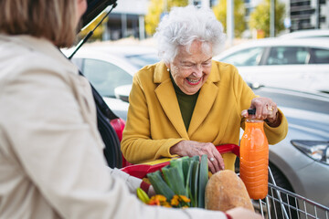 Mature granddaughter helping grandmother load groceries in to the car. Senior woman shopping at the shopping center. - obrazy, fototapety, plakaty