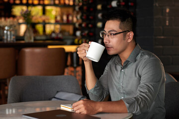 Pensive Vietnamese businessman drinking cup of coffee in restaurant during his lunch break