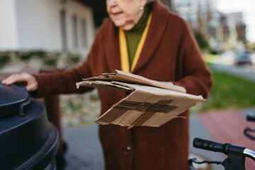 Elderly woman disposing cardboard into the garbage can, waste container in front of her apartment complex.