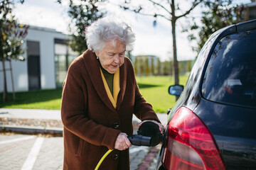 Close up of beautiful senior woman plugging charger in her electric car. Progressive elderly woman...
