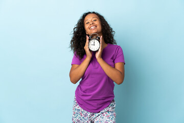 Teenager cuban girl isolated on blue background in pajamas and holding clock with happy expression