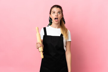 Young blonde woman holding a rolling pin isolated on pink background looking up and with surprised...
