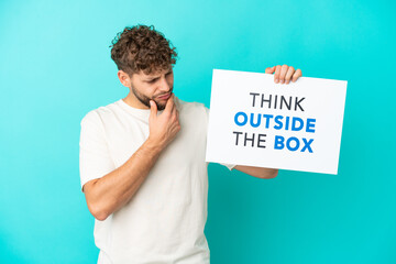 Young handsome caucasian man isolated on blue background holding a placard with text Think Outside The Box and thinking