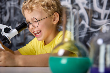 Young scientist of five years, squinting, attentively looks with one eye into the eyepiece of microscope, holding a tripod with his hand. Happy Caucasian boy in a yellow T-shirt studies a microscope - obrazy, fototapety, plakaty