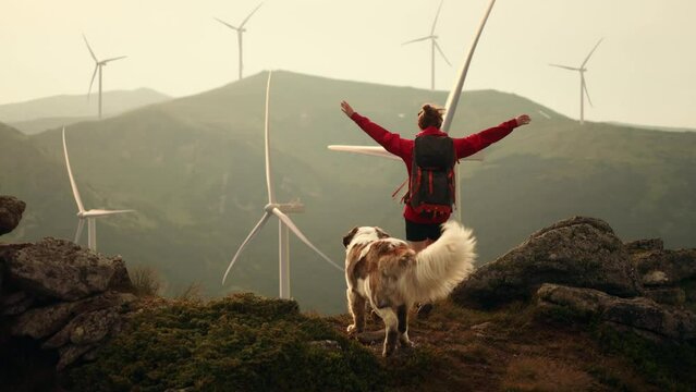 Young girl with saint Bernard dog on top of mountains raises arms into air and enjoying nature with wind turbine power station environmental engineering renewable energy on background
