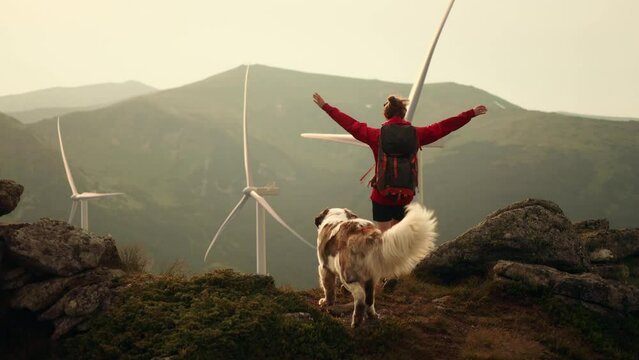 Young girl with saint Bernard dog on top of mountains raises arms into air and enjoying nature with wind turbine power station environmental engineering renewable energy on background