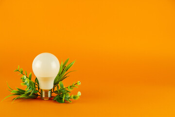 Energy saving light bulb and green leaves on orange background with copy space