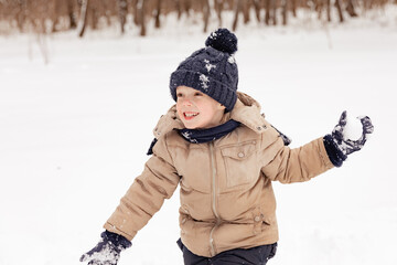 Fototapeta na wymiar Little boy having fun playing with fresh snow during snowfall. Snowball fight. Kid dressed in warm clothes, hat, hand gloves and scarf. Active outdoors leisure for child on nature in snowy winter day.