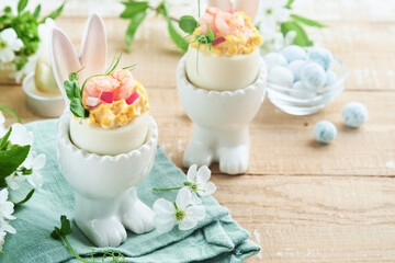 Obraz na płótnie Canvas Stuffed or deviled eggs with yolk, shrimp, pea microgreens with paprika in rabbit-shaped stand for easter table decorate fresh cherry or apple blossoms on light background. Traditional dish for Easter