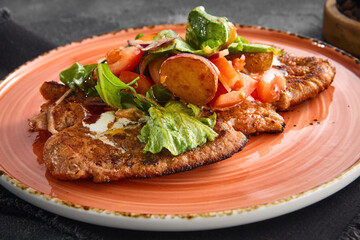 Breaded pork cutlets topped with a fresh and vibrant vegetable salad, artistically served on a salmon-colored ceramic plate, against a dark grey background