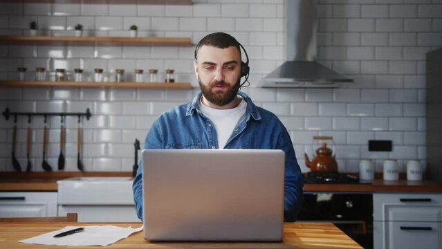 Bearded young adult business man wear wireless headset sitting against the kitchen counter having video conference calling on laptop computer talk by webcam in online chat 