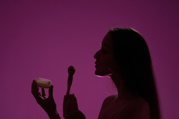 Side view of a dark silhouette of a woman holding a can of powder and a makeup brush in a studio on a pink background in pink neon light close up. Seminude woman blowing excess powder off makeup brush - obrazy, fototapety, plakaty