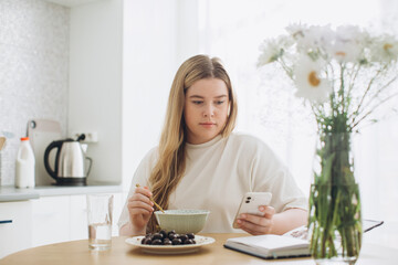 The blonde girl has breakfast while holding the phone in her hands.