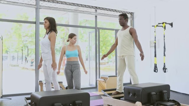 African-american man and latin woman doing exercises on reformers. Young caucasian woman pilates trainer correcting their moves. 