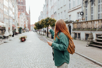 Attractive young female tourist is exploring city. Redhead girl with backpack pointing finger and...