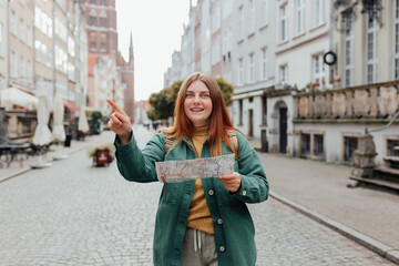 Attractive young female tourist is exploring city. Redhead girl with backpack pointing finger and holding a paper map on city street in Gdansk. Traveling Europe in autumn.